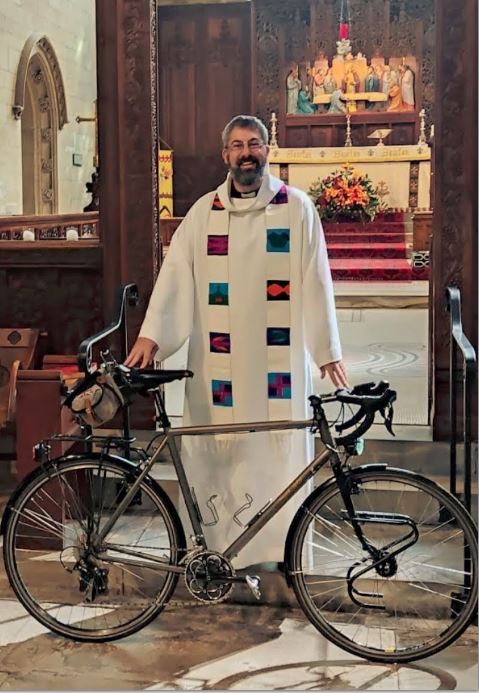 Vicar, a priest in the Church of England, standing in a church building with a bicycle in front of him. The priest is wearing robes.